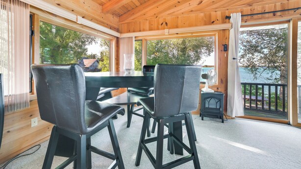 A wooden cabin with a dining area featuring two black chairs and a table, large windows with a view of trees and a lake, and a small heater on the floor. - Riverfront Sault Ste Marie Home w/ Dock! (Sault Ste. Marie)
