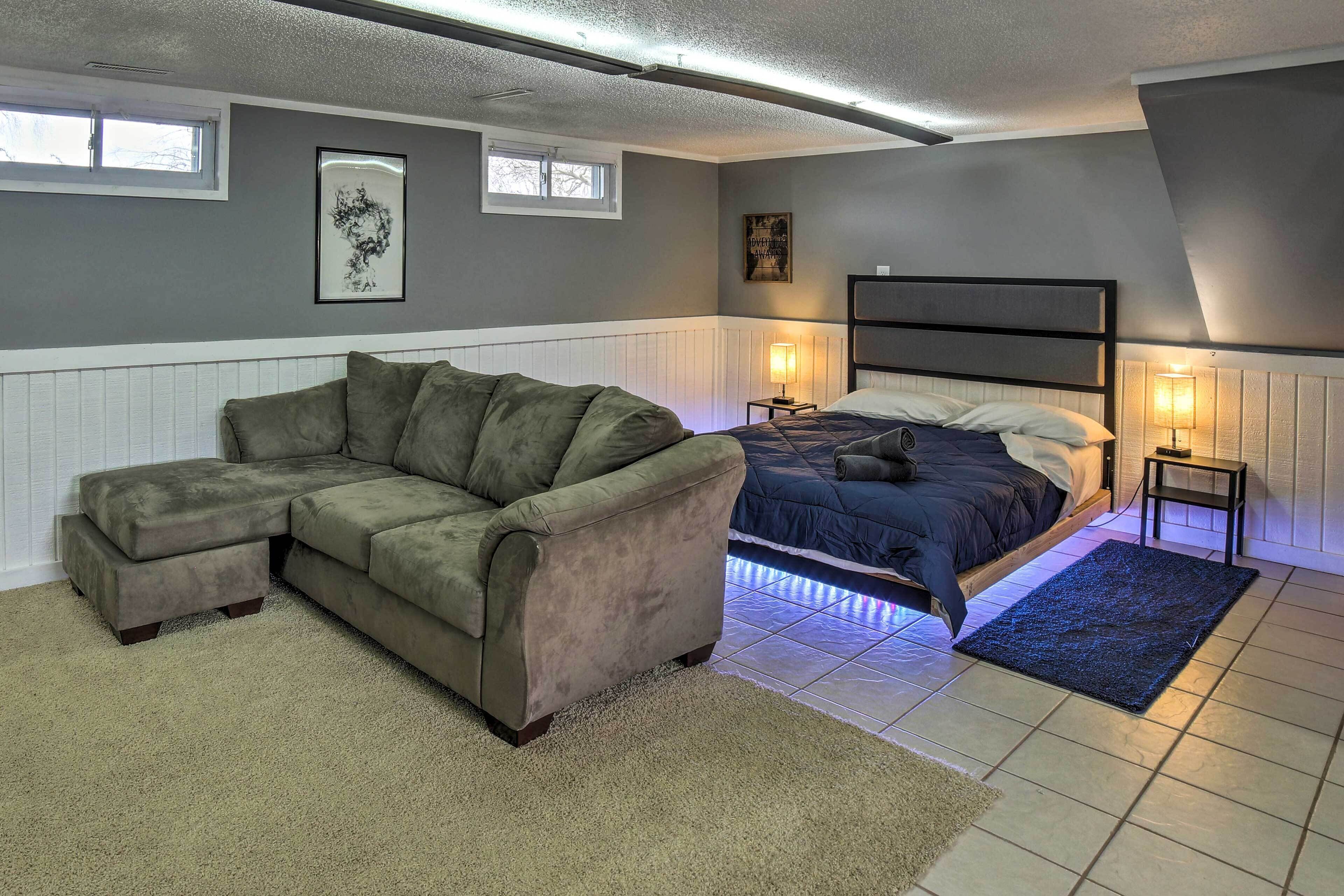 A bedroom with a sectional sofa, a bed with a headboard, two bedside tables with lamps, and a window.
