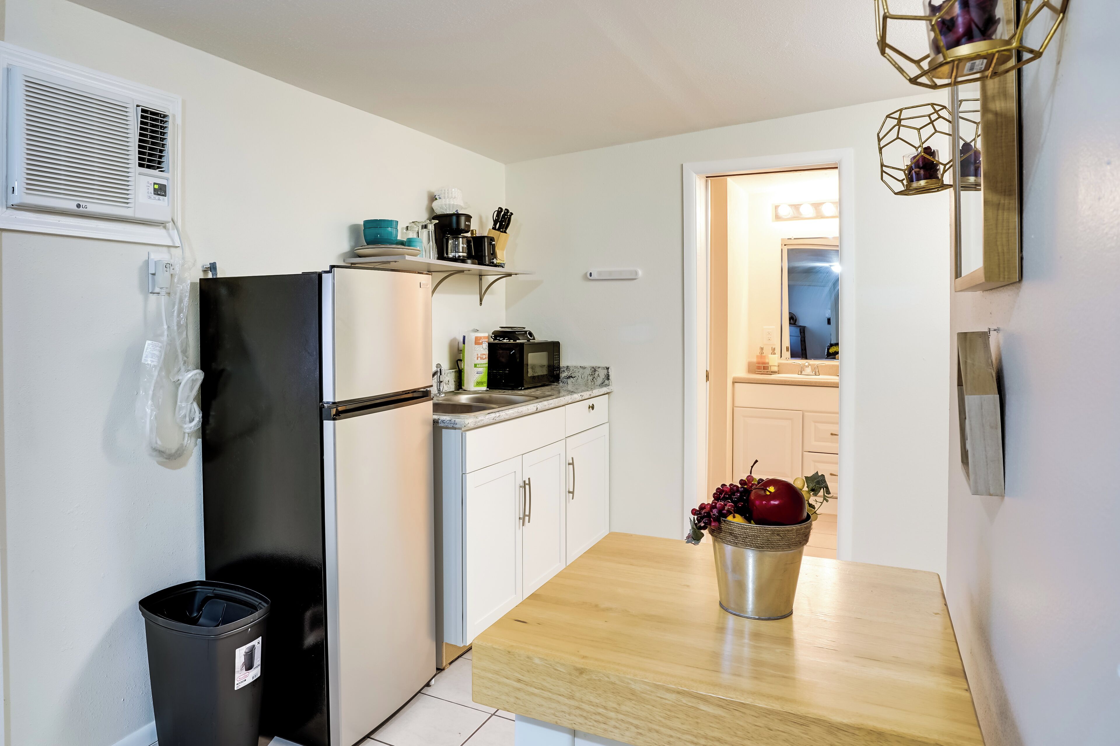 A compact kitchen with a refrigerator, microwave, and sink. A trash bin is visible to the left. A wooden table with a fruit bowl is in the foreground.