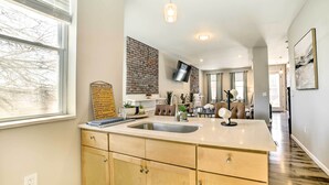 A modern kitchen with a large island, a brick accent wall, and a flat-screen TV mounted above the island. - Pittsburgh Townhome: 1 Mi to Downtown (Pittsburgh)