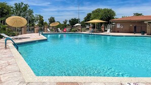 En klarblå pool med solstolar och parasoller, omgiven av en byggnad i tegel och träd.