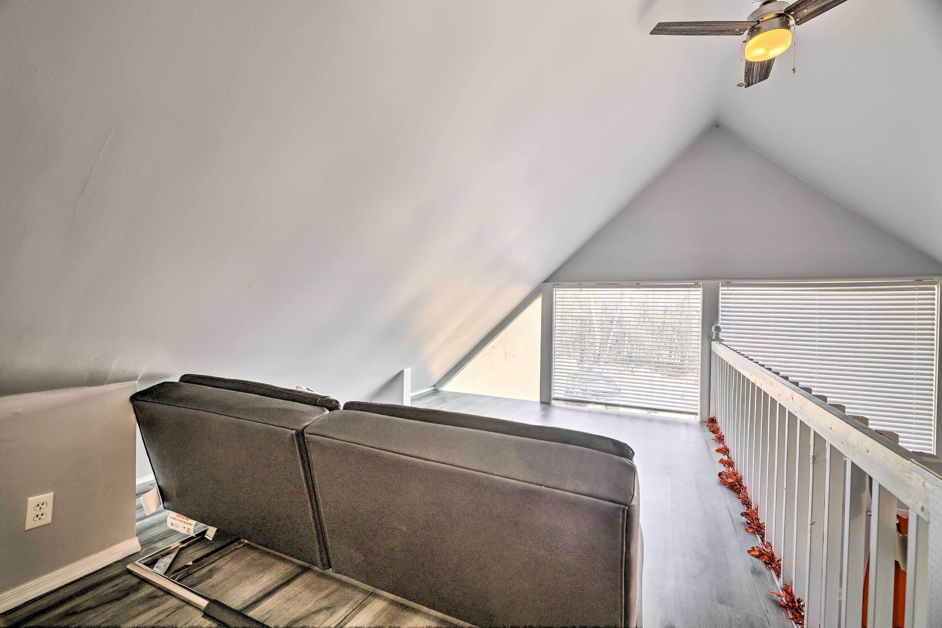 A room with a sloped ceiling, a ceiling fan, a window with blinds, and a white railing with red decorations.