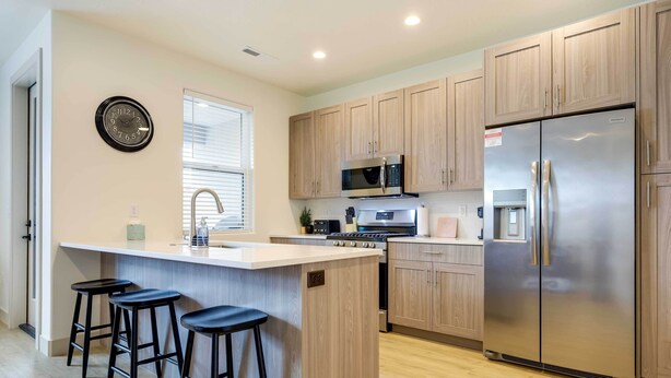 A modern kitchen with wooden cabinets, stainless steel appliances, a central island with stools, and a wall clock. - Modern Santa Clara Vacation Rental w/ Patio (Santa Clara)
