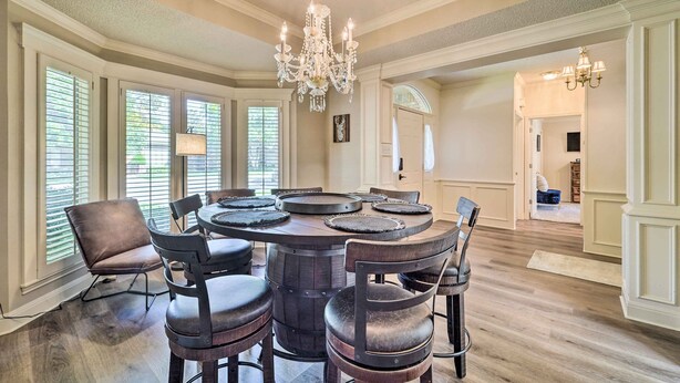 A dining area with a round table, six chairs, a chandelier, and large windows with blinds. - Lubbock Home w/ Patio ~ 6 Mi to Texas Tech! (Lubbock)