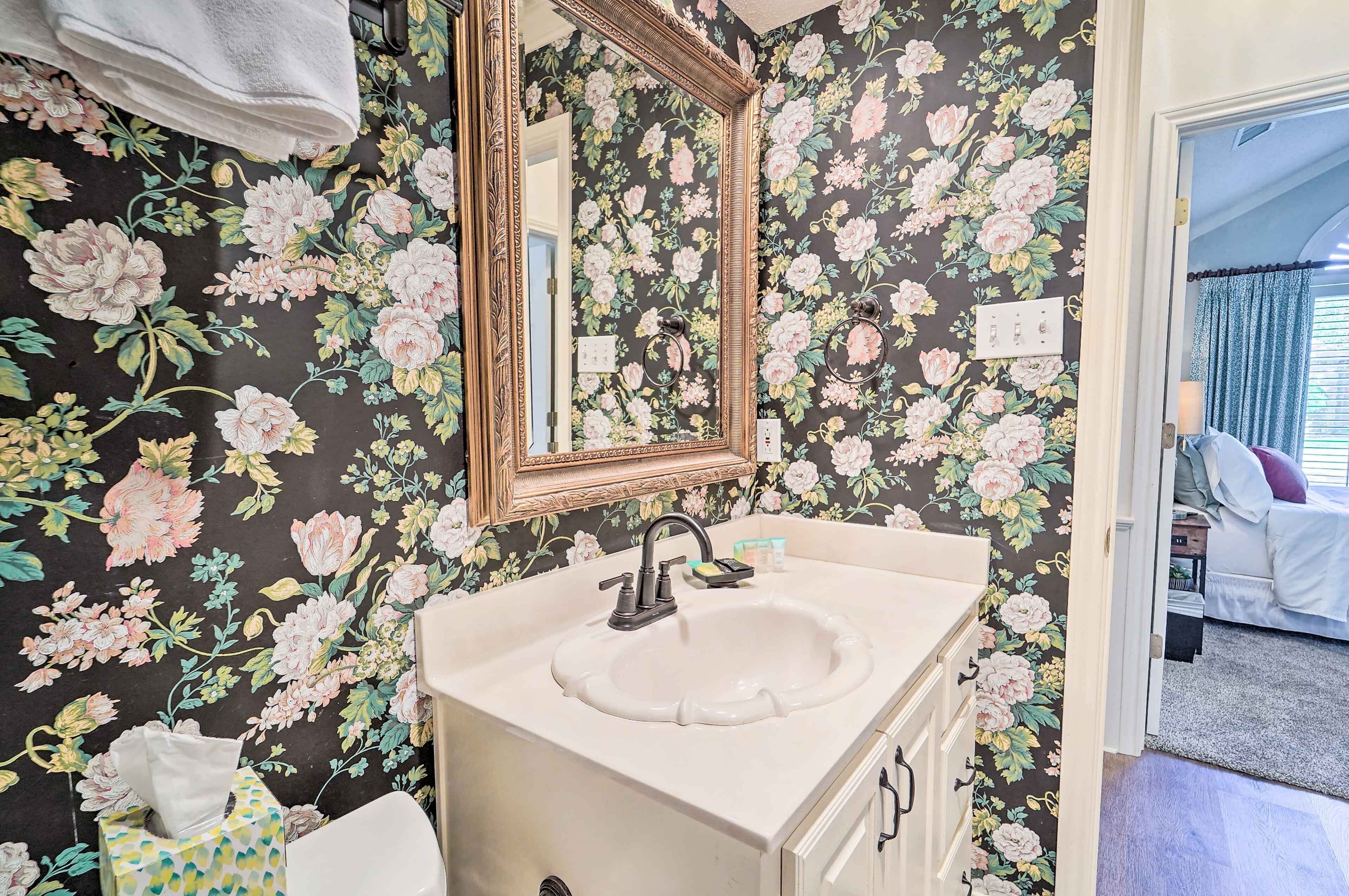 A bathroom with a floral wallpaper, a sink with a mirror, and a door leading to a bedroom.