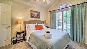 A neatly made bed with a green bedspread, a white pillow, and a red accent pillow. There is a nightstand with a lamp and a small table with a bowl of fruit. A painting hangs on the wall above the bed, and a window with curtains is visible. - Lubbock Home w/ Patio ~ 6 Mi to Texas Tech! (Lubbock)