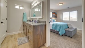 A bedroom with a bed, a gray ottoman, a window, and a bathroom with a sink and mirror. - Toquerville Vacation Rental, Near State Parks! (Toquerville)