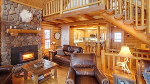 A log cabin interior with a stone fireplace, leather furniture, a wooden staircase, and a kitchen area. - Rustic Gotham Bay Cabin on Lake Coeur D'alene (Harrison)