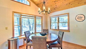 A dining area with a round table, six chairs, a chandelier, and large windows with a view of trees. - Pet-friendly Cabin in Pinetop-lakeside! (Pinetop)