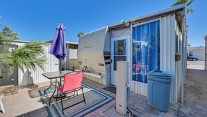 A compact RV with a sliding door, a small patio area with a table and chair, and a trash bin. - Cozy Yuma Vacation Rental w/ Resort Amenities! (Yuma)