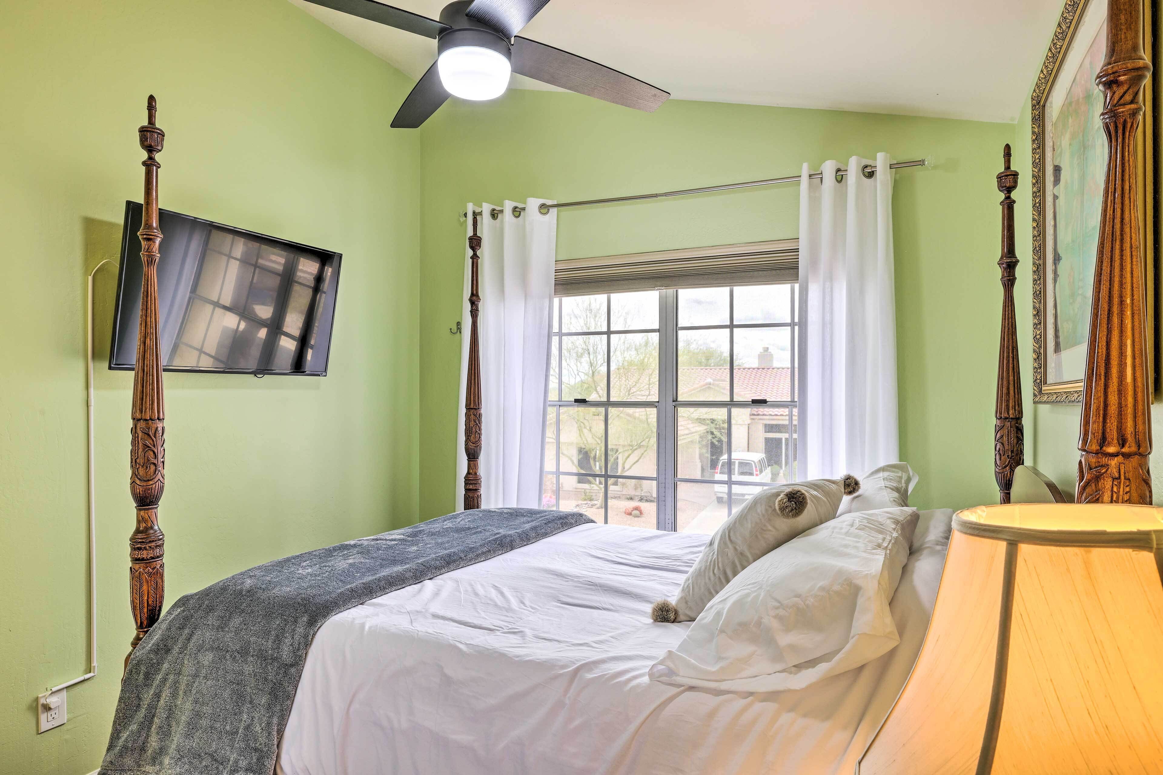 A bedroom with a bed, a ceiling fan, a window with curtains, and a television on the wall.