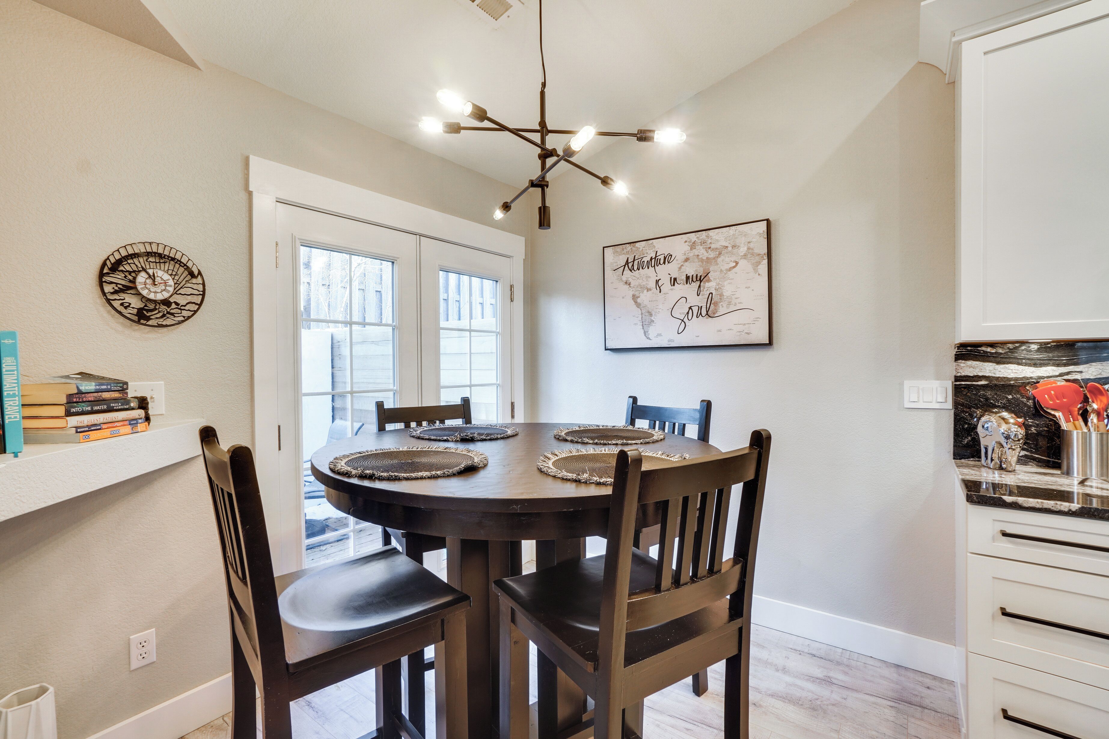 A dining area with a round table, four chairs, a chandelier, a wall clock, and a framed quote.
