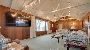 A cozy living room with wooden walls, a large flat-screen TV, floral-patterned sofas, and a ceiling with string lights. - Cozy Hillsdale Home ~ 1 Mi to Baw Beese Lake! (Hillsdale)