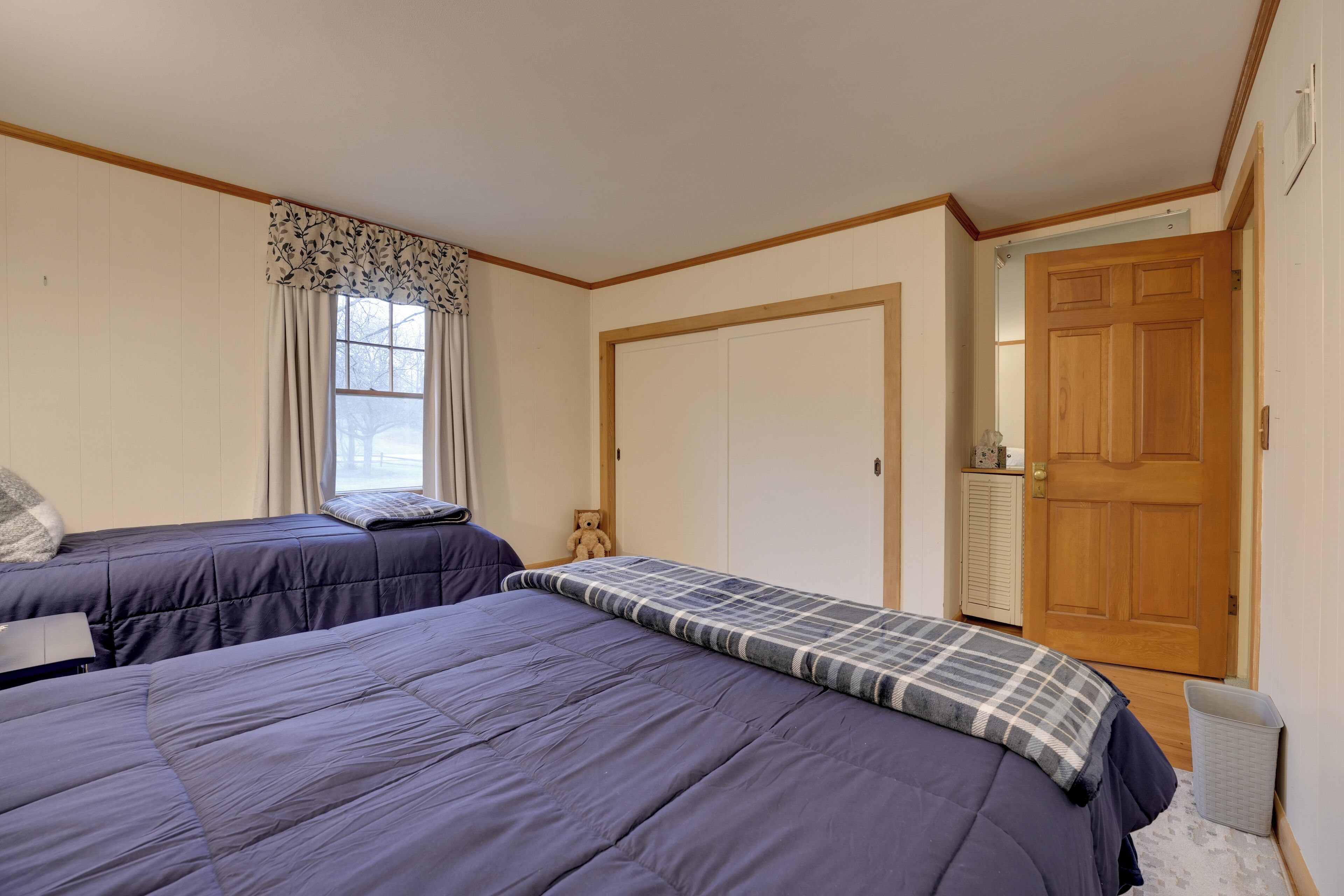 A bedroom with two beds, a window with curtains, a wooden door, and a small trash bin.