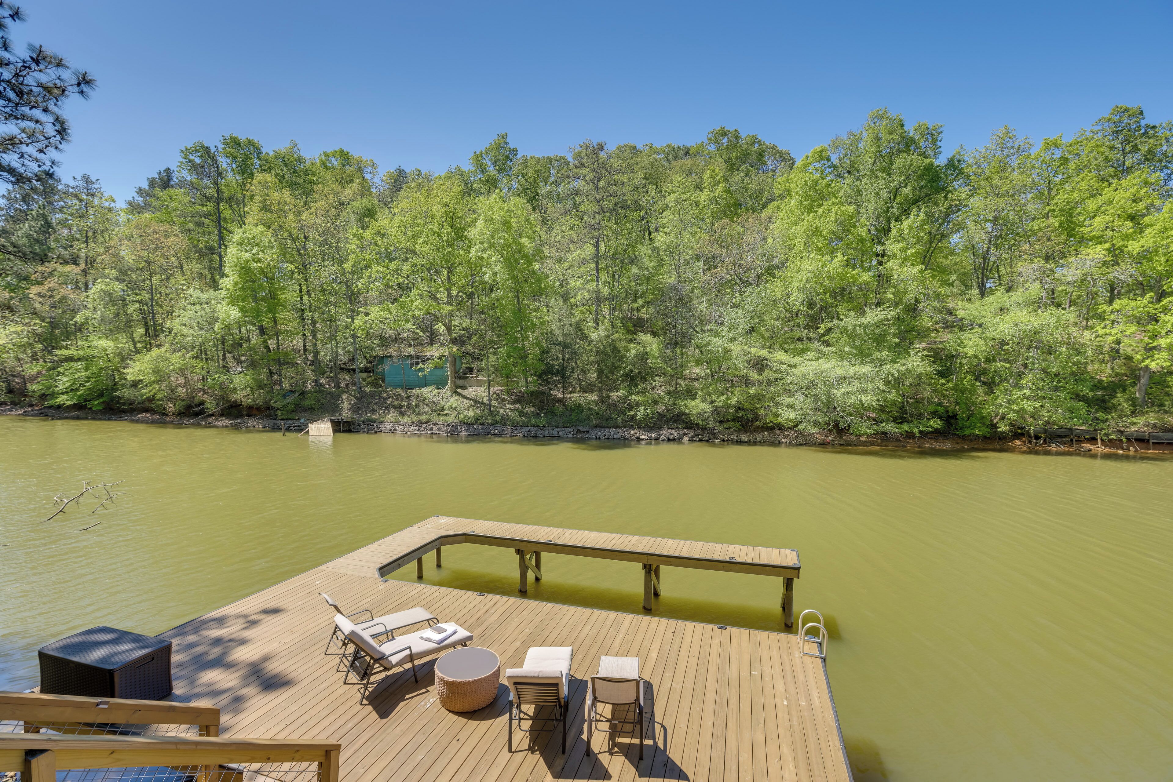 A wooden deck with lounge chairs and a table overlooking a calm body of water, surrounded by dense greenery.