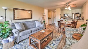 A living room with a grey sofa, a wooden coffee table, and a patterned rug. There is a dining area with a table and chairs, and a kitchen in the background. - 'deja Blue' Oceanside Condo on Beach w/ Pools! (Oceanside)