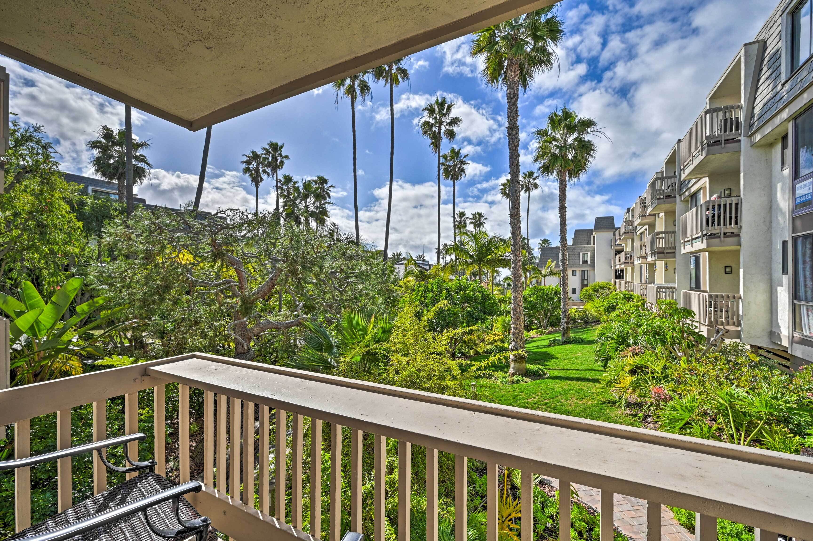 A balcony with a view of a lush garden, tall palm trees, and a row of residential buildings.
