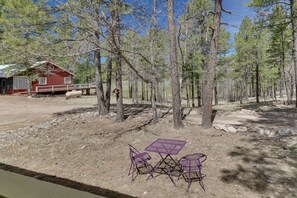 A rustic cabin with a red exterior, surrounded by a forest. There is a small table and two chairs in the foreground. - Pet-friendly Alpine Cabin w/ Gas Grill & Fireplace (Alpine)
