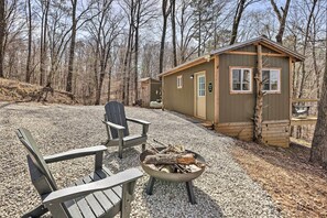 A cabin with a gravel driveway, two Adirondack chairs, and a fire pit with logs. - Cozy Studio Cabin in Tallassee w/ Water View! (Tallassee)