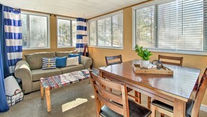 A cozy living room with a sofa, a wooden bench, a dining table with chairs, and large windows with blinds.