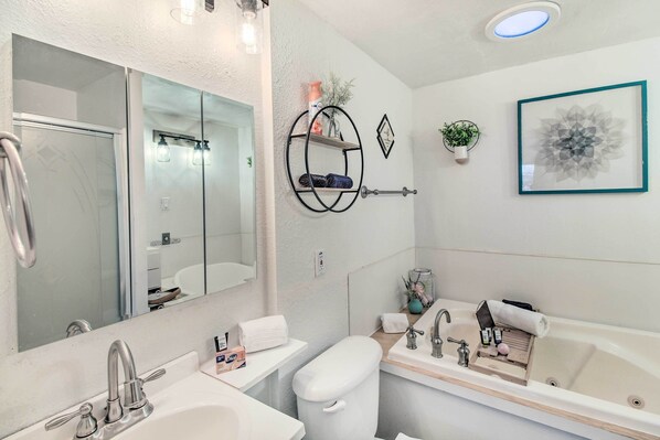 A modern bathroom with a large mirror, a toilet, a sink, and a bathtub. There are shelves with toiletries and a framed artwork on the wall.