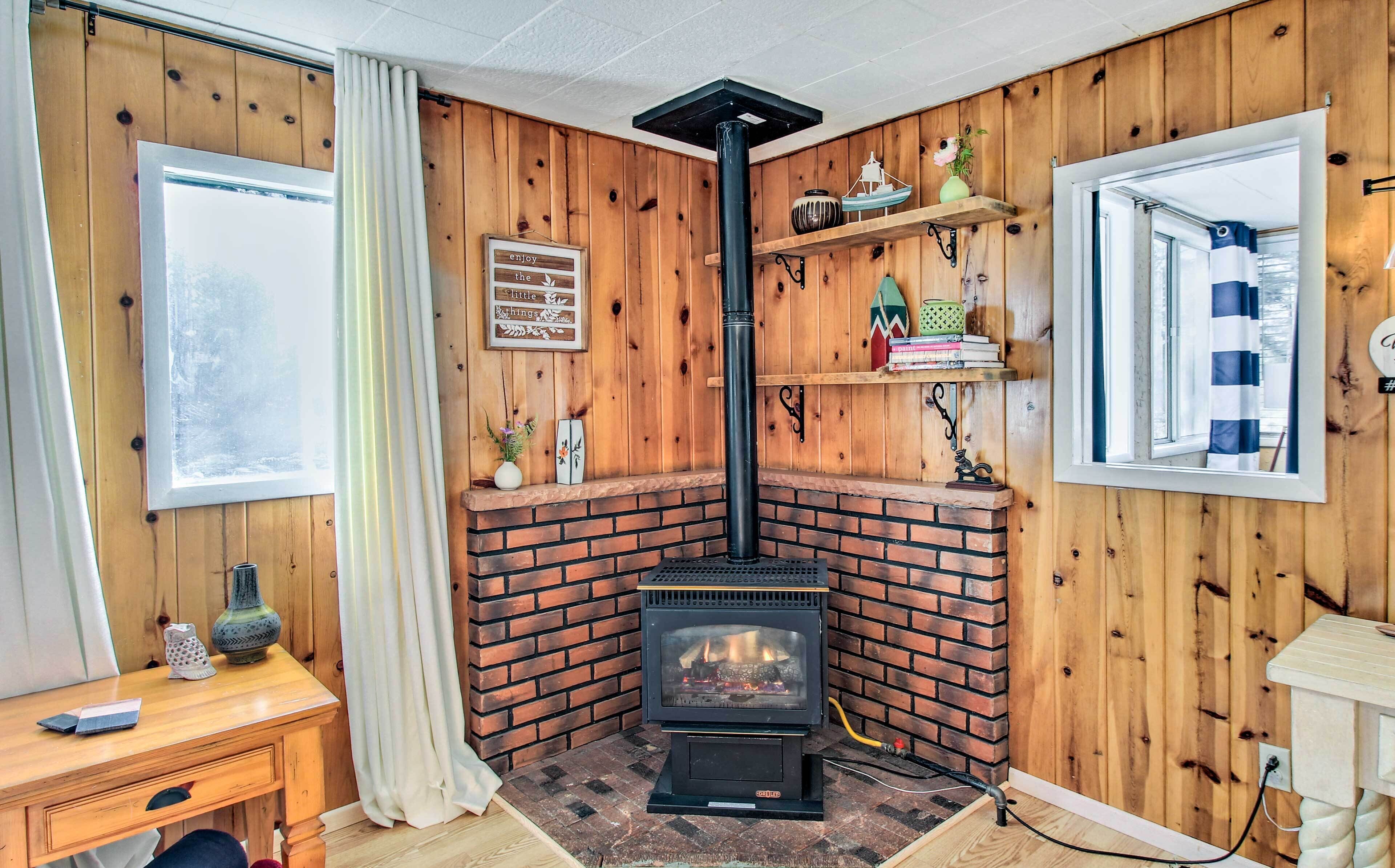 A cozy room with a brick fireplace, wooden walls, a window with curtains, and shelves with decorative items.