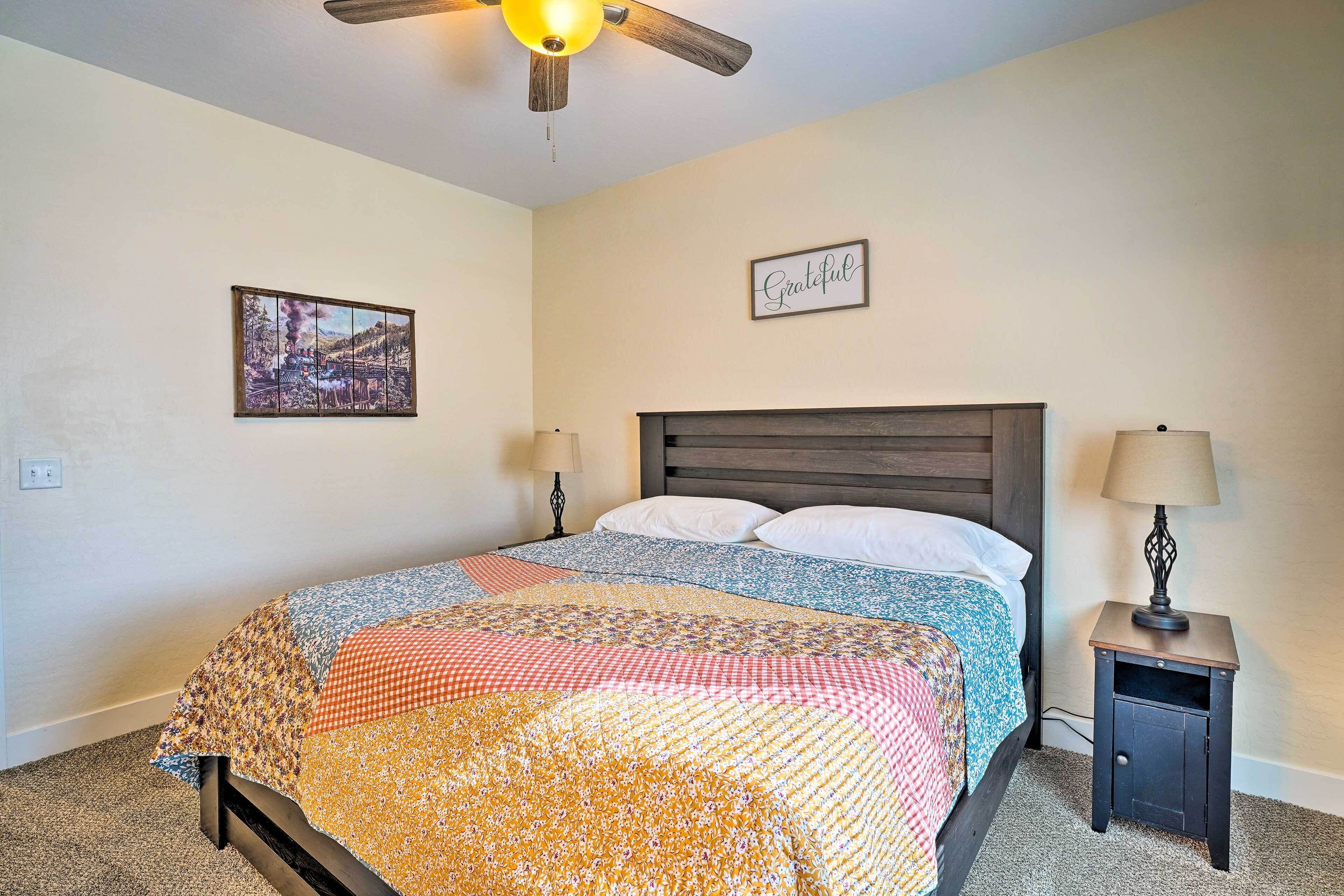 A bedroom with a bed, a nightstand, a wall-mounted lamp, a ceiling fan, a framed picture, and a 'Grateful' sign.