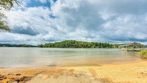 A beach with a wooden pier extending into calm waters, surrounded by lush greenery and a cloudy sky. - Cozy Lake Chatuge Getaway, Walk to Water! (Hayesville)