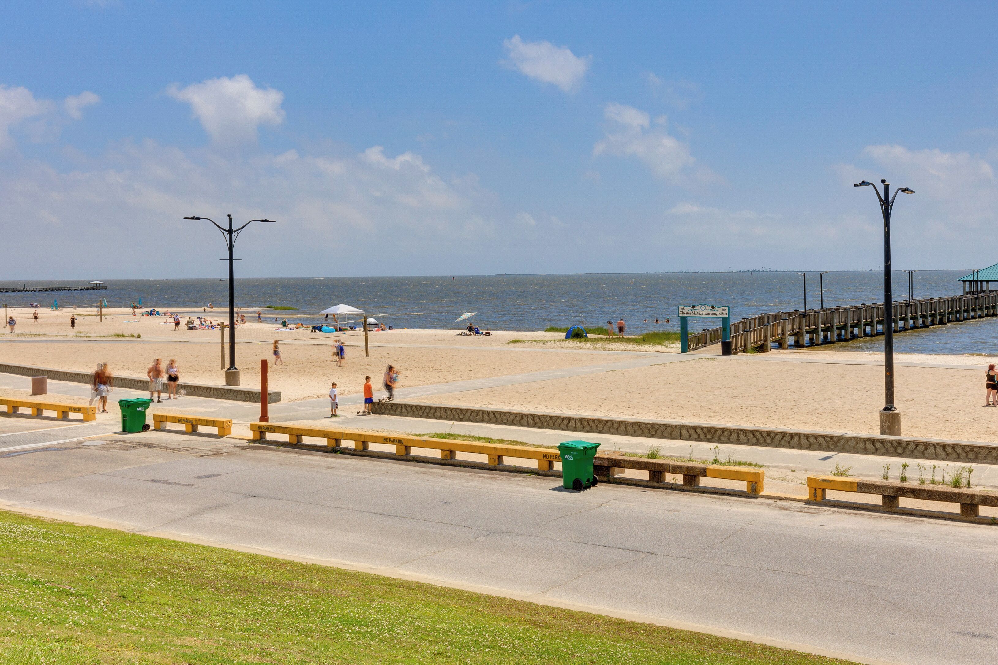 A beach with a wooden pier, a road with yellow barriers, and a green trash bin.