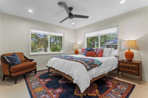 Quarto com cama, duas mesinhas de cabeceira, uma cadeira, ventoinha no teto e janelas amplas.