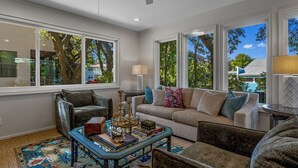 Ein Wohnzimmer mit einer Couch, Sesseln, einem Couchtisch und großen Fenstern mit Blick auf Bäume und ein Haus.
