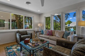 A living room with a sofa, chairs, a coffee table, and large windows with a view of trees and a house. - Live Oaks & Marina Views! Ocean Springs Home (Ocean Springs)