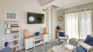Ein Wohnzimmer mit an der Wand montiertem Fernseher, einem Bücherregal, einem Couchtisch und einer Sitzecke mit gepolstertem Sitz und Stühlen.
