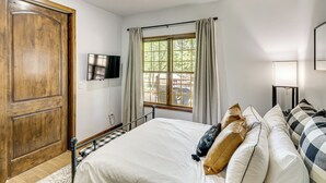 Un dormitorio con una puerta de madera grande, una cama con muchos almohadones, una ventana con cortinas y un televisor de pantalla plana montado en la pared.