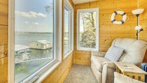 En mysig stuga med stora fönster som vetter mot en brygga och vattnet, en bekväm soffa och ett litet bord med en lampa.