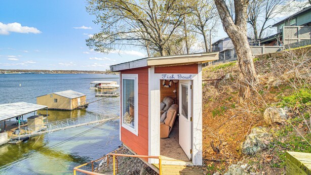 En liten, envåningsstuga med en skylt som visar en fiskeskål, belägen nära en brygga med båtar, omgiven av träd och en sjö eller ett vattendrag.