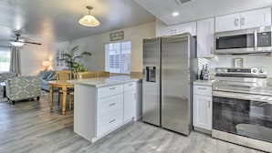 Una cocina moderna con armarios blancos, electrodomésticos de acero inoxidable, una zona de comedor con mesa y sillas de madera, y un ventilador de techo.