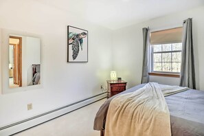 A bedroom with a bed, a bedside table with a lamp, a window with curtains, a wall-mounted picture, and a door leading to another room.