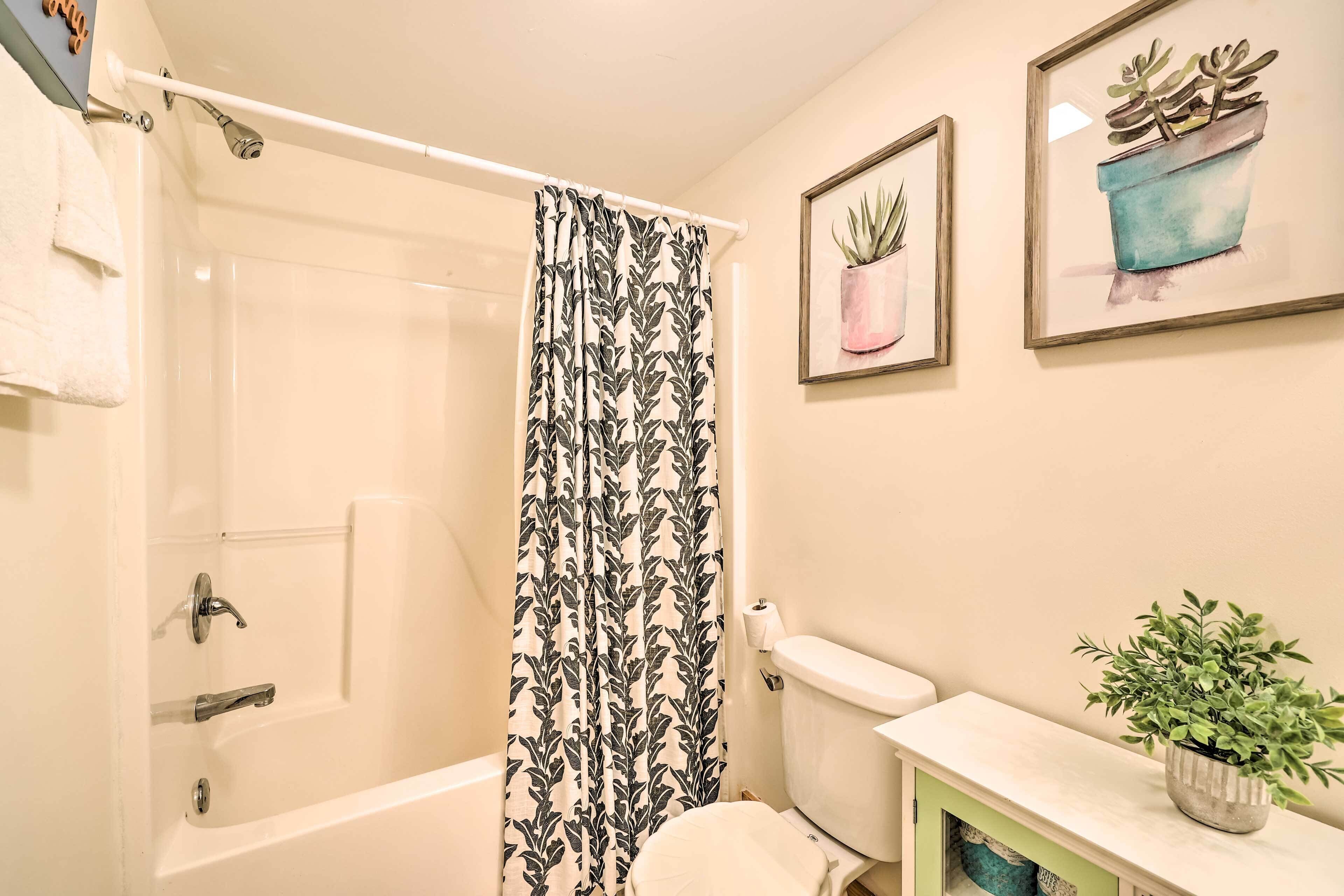A bathroom with a bathtub, toilet, and a shower curtain with a leaf pattern. There are two framed pictures on the wall, one of a plant in a pot and another of a plant in a pot.