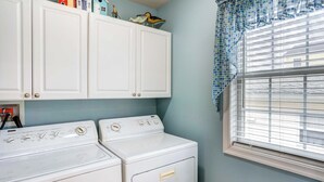 A laundry area with a front-loading washer and dryer, white cabinets above, and a window with blinds. - Bowers Beach Vacation Rental: 2 Blocks Away! (Frederica)