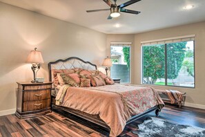 A bedroom with a bed, wooden nightstand, two bedside lamps, a ceiling fan, and a large window with a view of greenery. - Phoenix Home: Heated Pool, Patio & Mountain Views! (Phoenix)