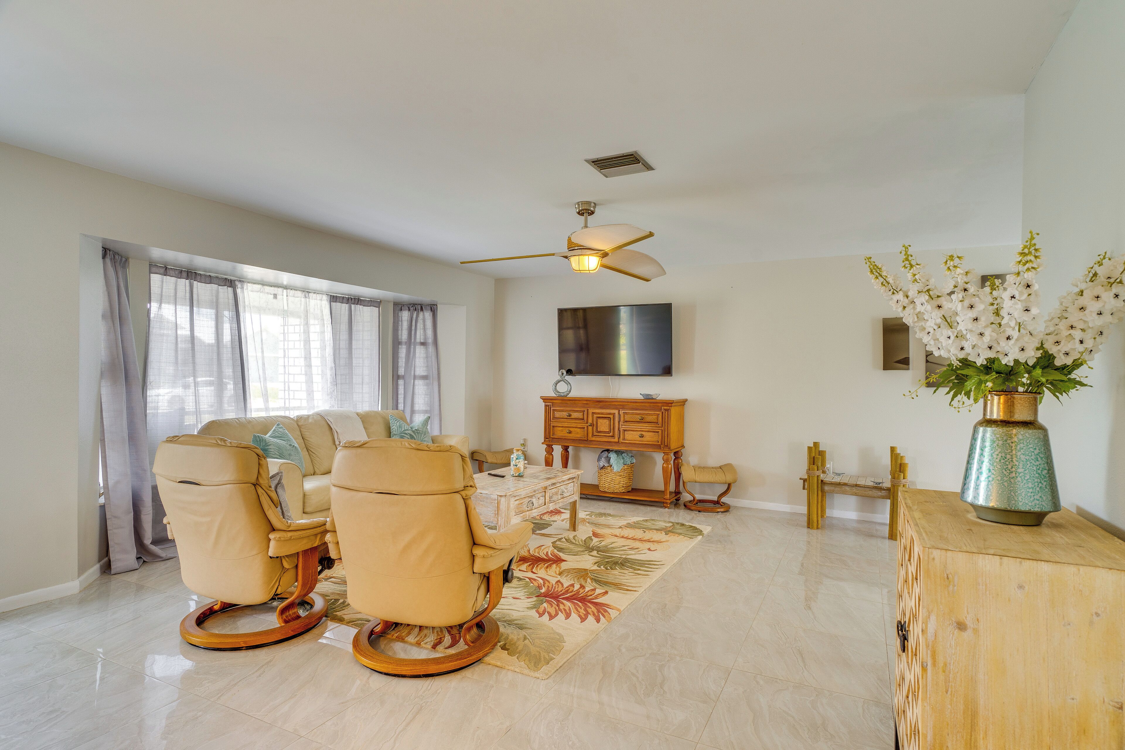 A spacious living room with a beige sofa, two rocking chairs, a wooden coffee table, and a flat-screen TV mounted on the wall.