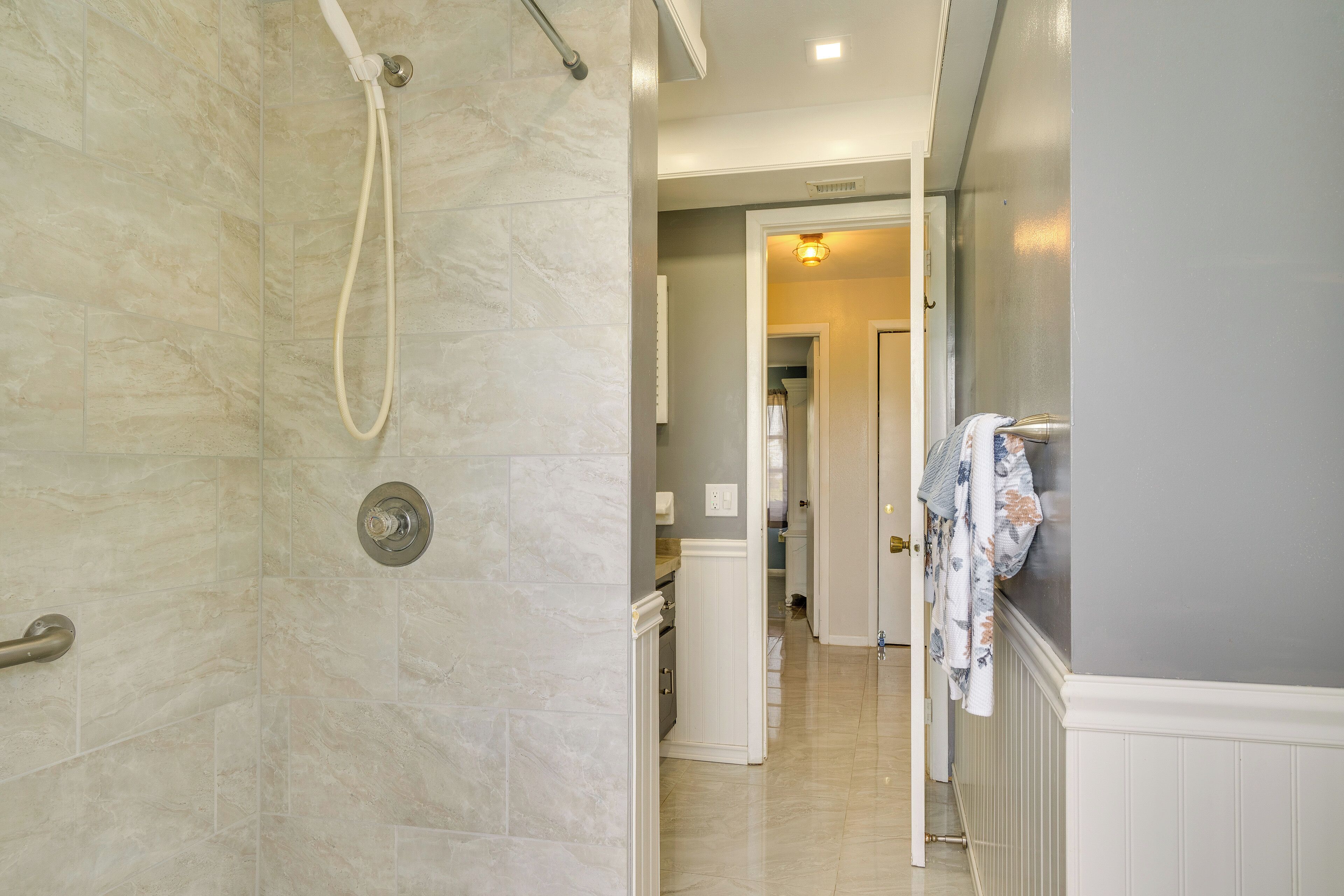 A bathroom with a shower, a toilet, and a sink. There is a hallway leading to a kitchen area.