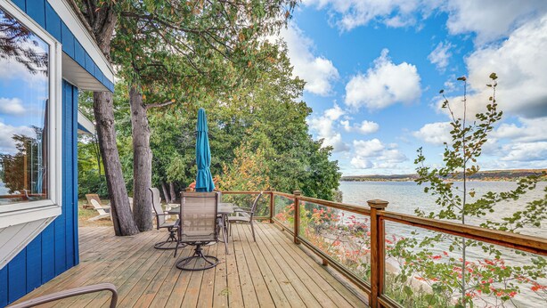 Eine Holzterrasse mit Gartenmöbeln, ein blaues Gebäude und ein See im Hintergrund.