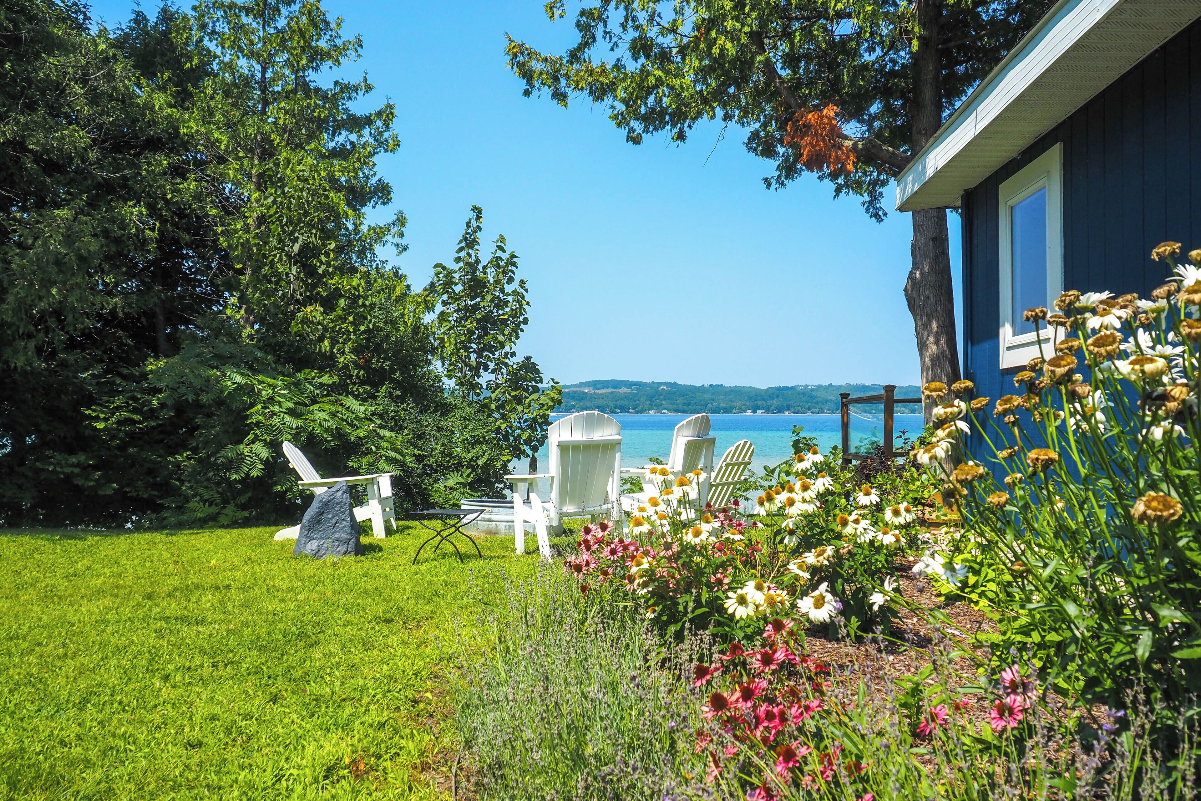A blue house with a view of a lake, surrounded by a grassy lawn and white Adirondack chairs.