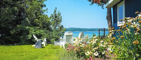 A blue house with a view of a lake, surrounded by a grassy lawn and white Adirondack chairs.