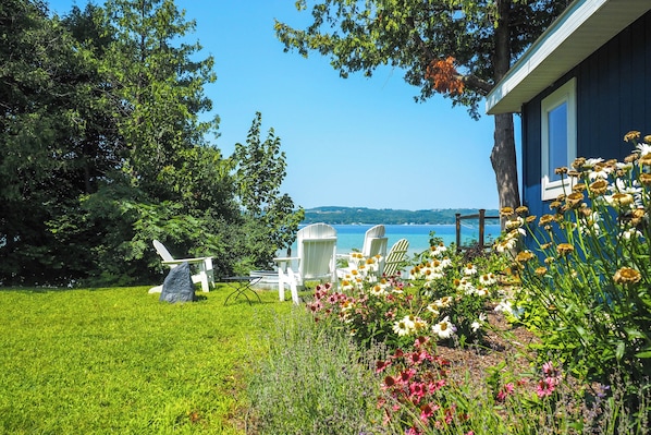 A blue house with a view of a lake, surrounded by a grassy lawn and white Adirondack chairs.