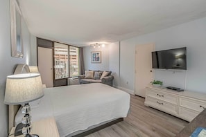 A bedroom with a bed, a nightstand, a lamp, a dresser, a TV, and a window with a view of buildings. - Balcony & Resort Amenities at Myrtle Beach Condo! (Myrtle Beach)