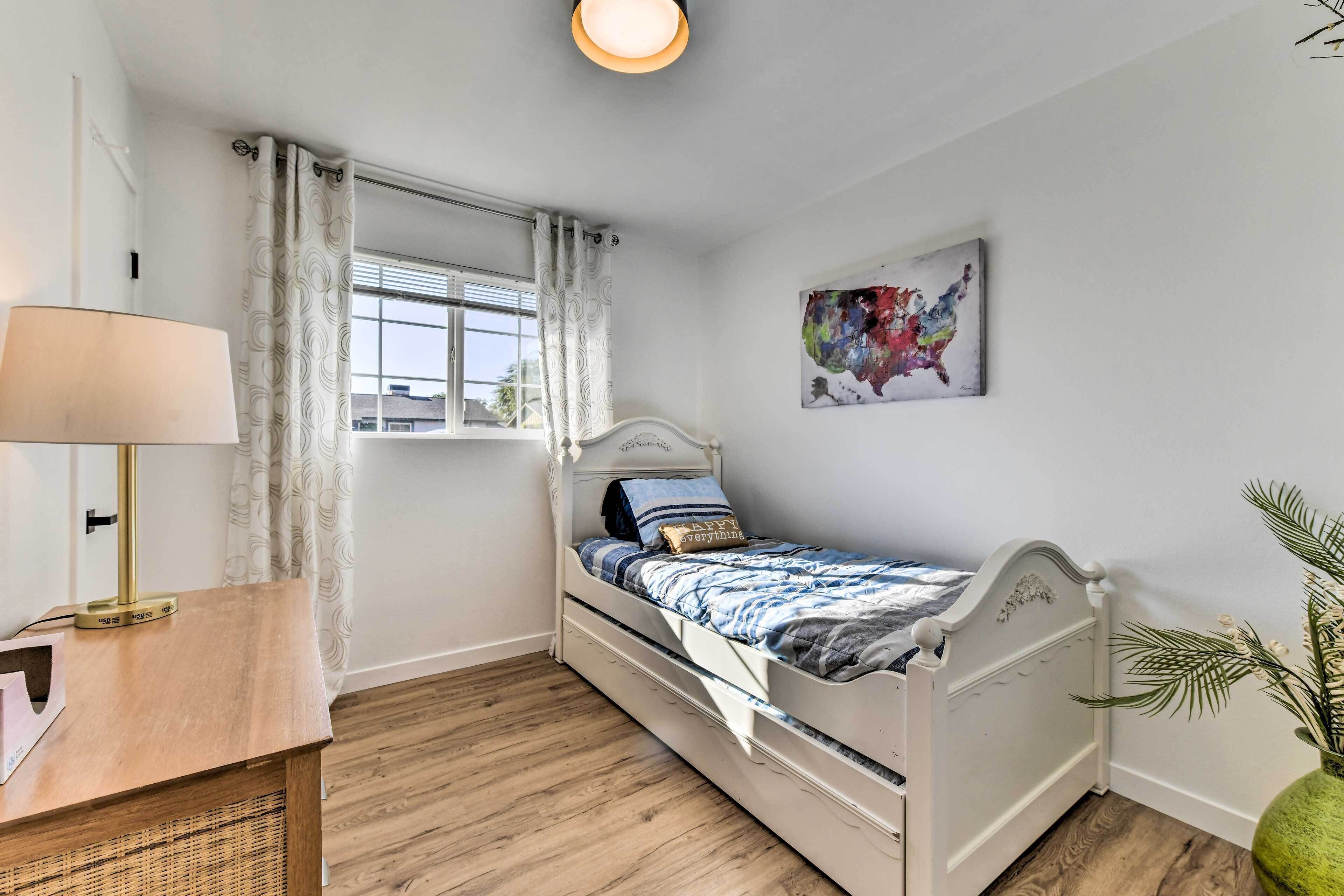 Un dormitorio con cama, mesita de noche, lámpara, ventana con cortinas, un cuadro colgado en la pared y una planta en maceta.