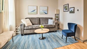 A living room with a grey sofa, a blue armchair, a round wooden coffee table with a vase of flowers, and a striped rug. - Cozy Omaha Vacation Rental: 6 Miles to Downtown! (Omaha)